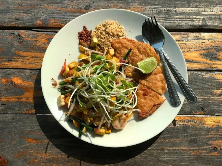 Assiette de spécialités thaïlandaises garnie de légumes, d'herbes fraîches et de différents ingrédients, photographiée sur une table en bois.