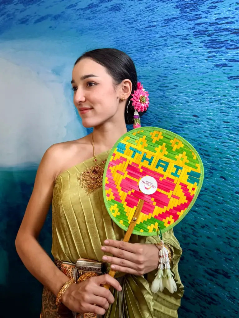 Jeune femme vêtue d'une tenue traditionnelle thaïlandaise dorée tenant un éventail tressé multicolore.