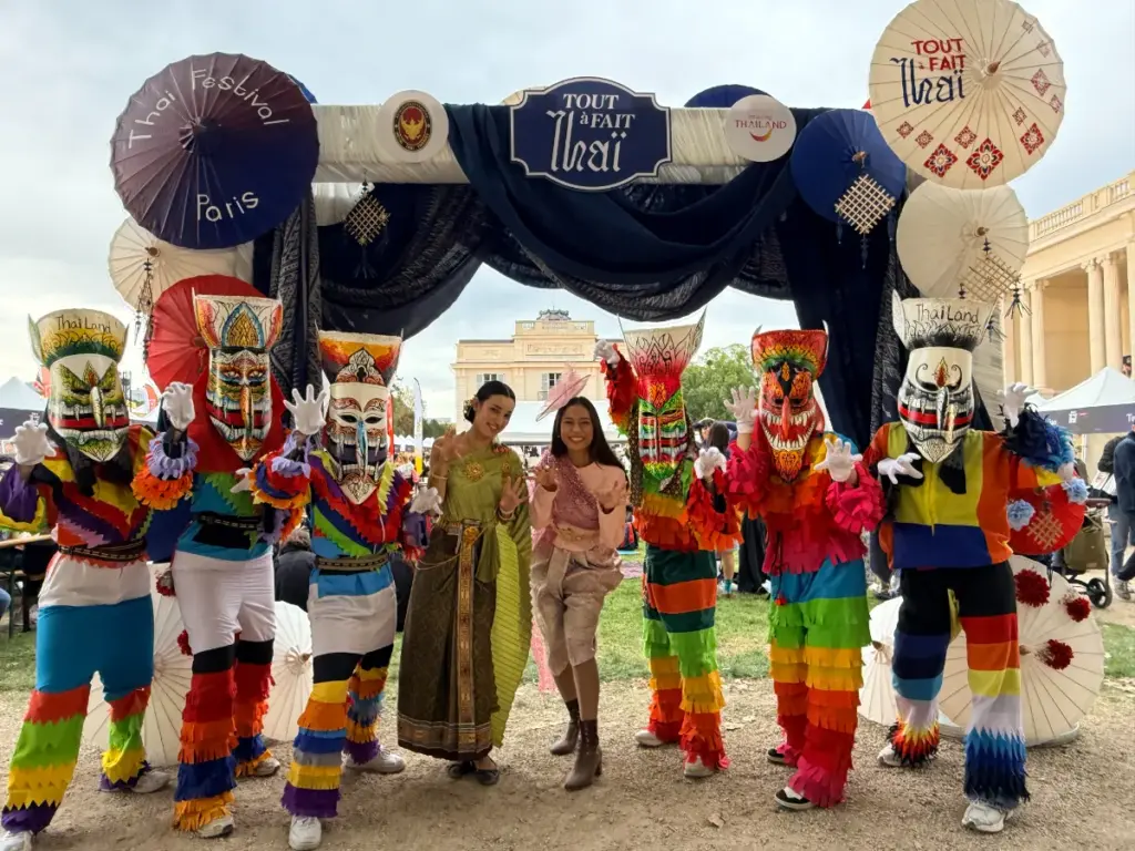 Deux femmes en tenues traditionnelles thaïlandaises posent aux côtés de personnages portant de grands masques colorés.
