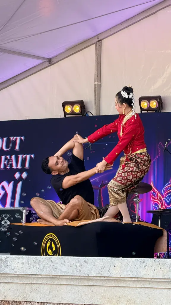 Une praticienne en tenue traditionnelle thaïlandaise rouge réalise une démonstration de massage et d'étirement sur un participant assis, sur la scène du festival Tout à fait Thaï 2026.