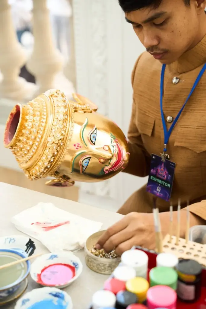 Artisan tenant un masque traditionnel thaïlandais doré, orné de motifs colorés, lors d'une présentation du savoir-faire artisanal thaïlandais.