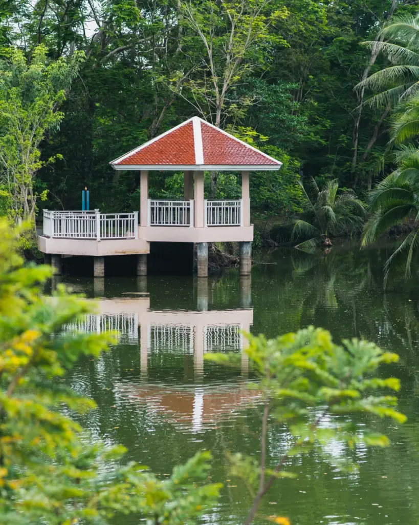Petit pavillon à deux niveaux avec un toit rouge, entouré d'une végétation tropicale dense et se reflétant dans l'eau.